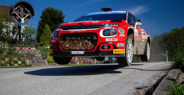 Ein rotes Rallye-Auto, das über eine Straßenkuppe springt - im Hintergrund ist ein Bildstock mit der Darstellung des gekreuzigten Jesus zu sehen