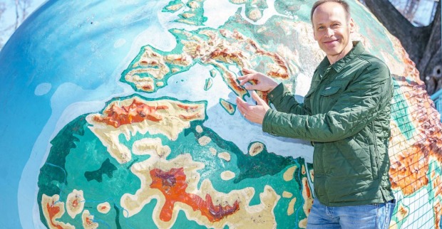 Meteorologe Marcus Wadsak steht vor einer Weltkugel und zeigt mit dem Finger auf ein Land