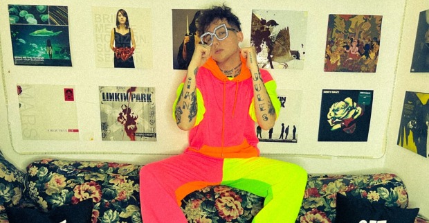 Ian Jules, der in einem neonfarbenen Outfit auf der Rückenlehne einer Couch sitzt. Im Hintergrund sind zahlreiche Plattencover an der Wand zu sehen.