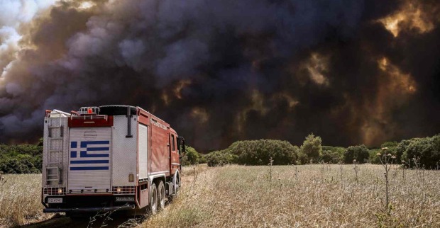 Ein Feuerwehrauto fähr über eine völlig vertrocknete Wiese auf eine Schwarze Wand aus Rauch und Flammen zu.