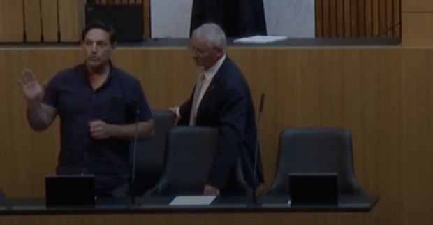 Der Eindringling steht bei der Regierungsbank und redet mit erhobener Hand. Ein Mann in Anzug fasst ihn von der Seite an.