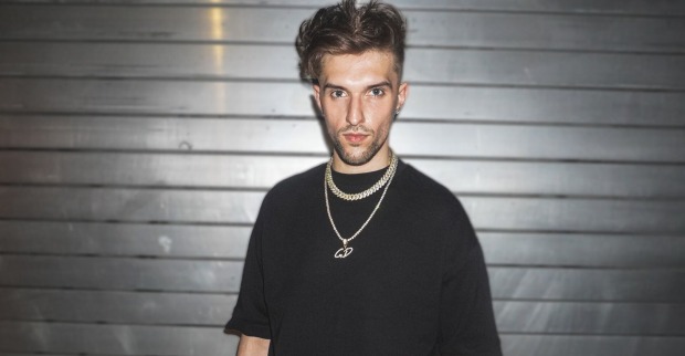 Gavis Dean, der ein schwarzes T-Shirt und eine Goldkette trägt, und vor einem Rolltor steht