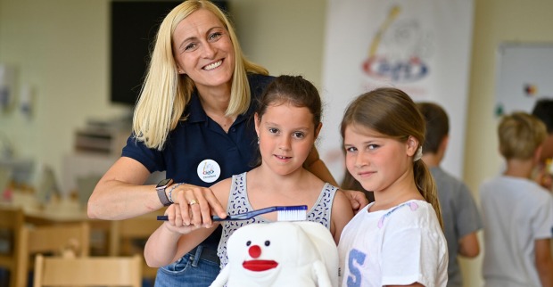 Eine Frau zeigt zwei Mädchen, wie sie richtig Zähne putzen anhand eines Maskottchens (Backenzahn).