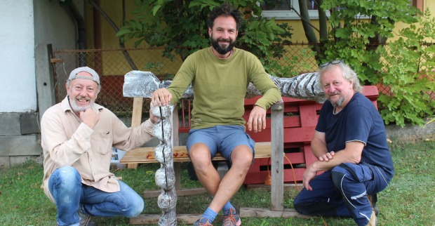 Drei Bildhauer, sitzend und kniend, lächeln in die Kamera.