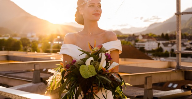 Junge Frau steht bei Sonnenuntergang im Brautkleid mit Blumen in den Händen