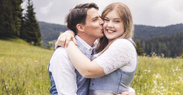 Ein Mann in Tracht gibt einer Frau im Dirndl mit blonden Haaren einen Kuss auf die Wange. Sie stehen auf einer Wiese.