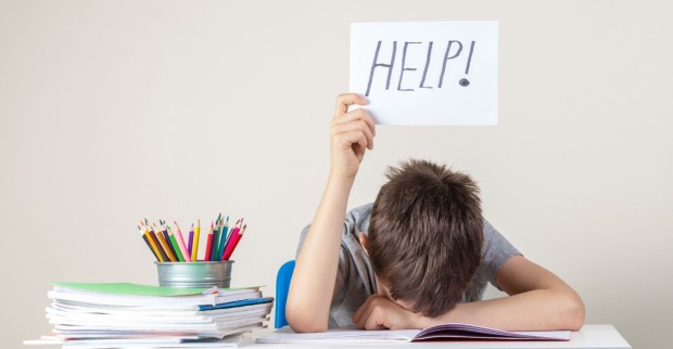 Ein junger Schüler stützt seinen Kopf auf seinen Unterarm auf einem Schreibtisch auf und hält ein Schild mit der Aufschrift "Help!" in die Höhe