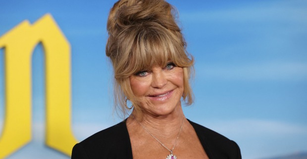 Schauspielerin Goldie Hawn trägt hochgesteckte Haare und steht vor einer blauen Leinwand.