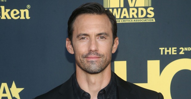 Schauspieler Milo Ventimiglia vor einer schwarzen Fotowand mit einem schwarzen Anzug und gegolten Haaren.