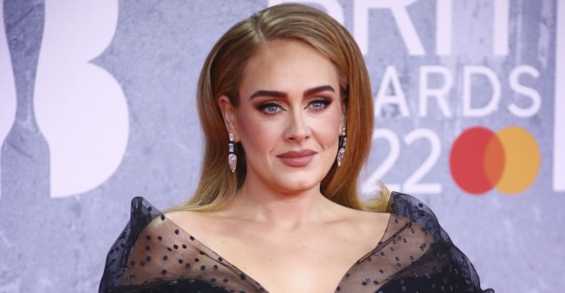 Adele trägt ein schwarzes Kleid mit Spitze und einen tiefen Seitenscheitel. Die Haare sind dunkelblond und offen. Sie steht vor einer violetten Fotowand.