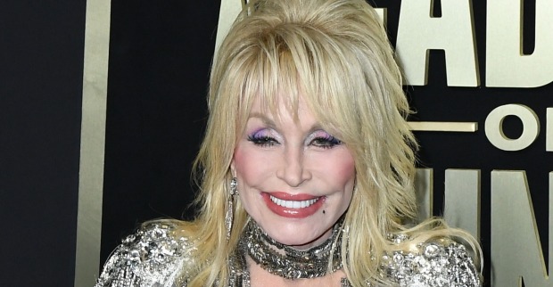 Dolly Parton hat blonde, toupierte Haare, viel Make-up und ein glitzerndes Kleid. Sie steht vor einer schwarzen Fotowand.