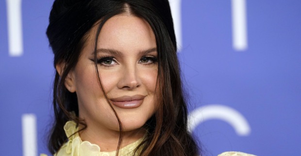 Lana Del Rey trägt hochgesteckte braune Haare und ein starkes Augen Make-Up. Sie steht vor einer blauen Wand.