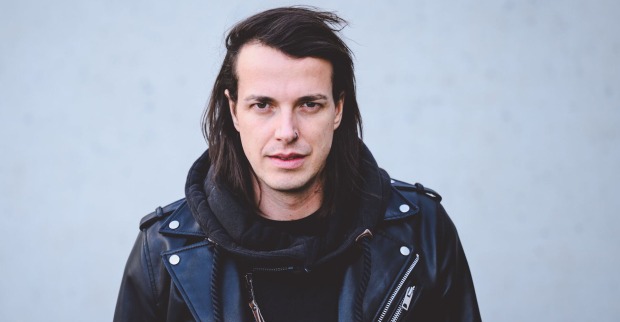 Marco Pogo trägt einen schwarzen Pullover und eine schwarze Lederjacke. Er schaut frontal in die Kamera und steht vor einer grauen Wand.