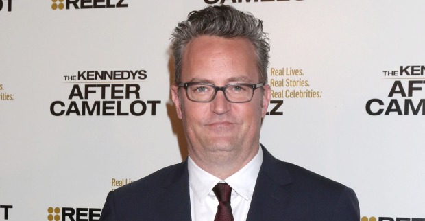 Matthew Perry trägt ein schwarzes Sakko und eine weiße Bluse und eine Brille. Er steht vor einer Fotoleinwand.