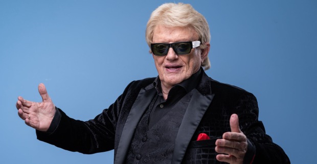 Heino steht vor einer blauen Fotowand mit einem schwarzen Sakko, blonden Haaren und einer Sonnenbrille.