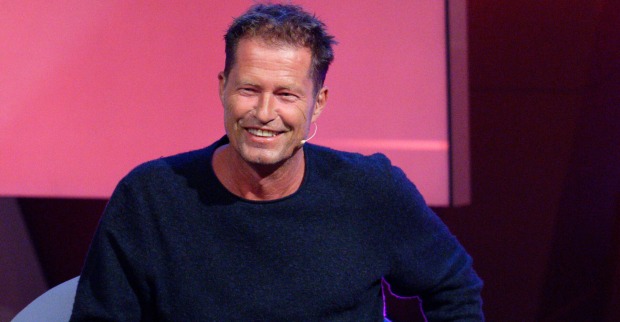 Til Schweiger mit einem dunkelblauen Pullover vor einer Rosinen Filmwand.