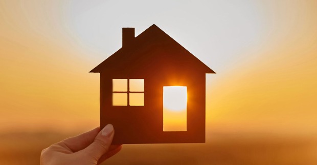 Junge Frau hält ein Miniaturhaus gegen das Licht der untergehenden Sonne | Credit: iStock.com/bodnarchuk