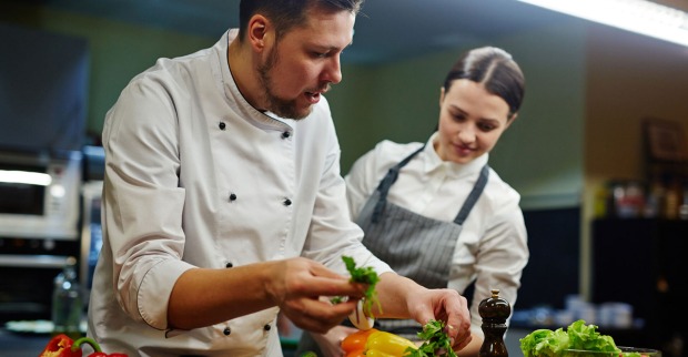 Koch bringt einem weiblichen Lehrling bei, wie man ein Gericht appetitlich anrichtet | Credit: iStock.com/shironosov