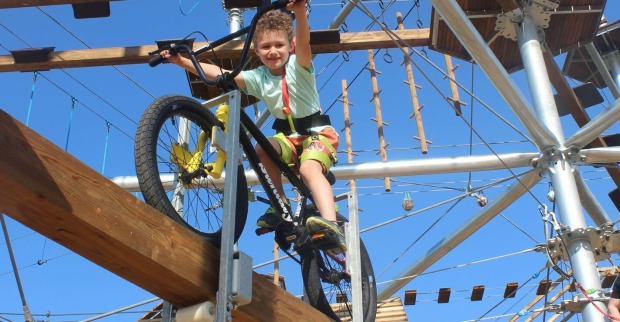 Ein junge balanciert mit einem Mountainbike auf einem Holzbalken. Er trägt ein grünes T-Shirt und eine kurze, gelbe Hose. Er ist mit Seilen und einem Gurt gesichert. Im Hintergrund sieht man den Klettergarten des Naturparks Ikuna mit Brücken, Seilen und anderen Hindernissen.