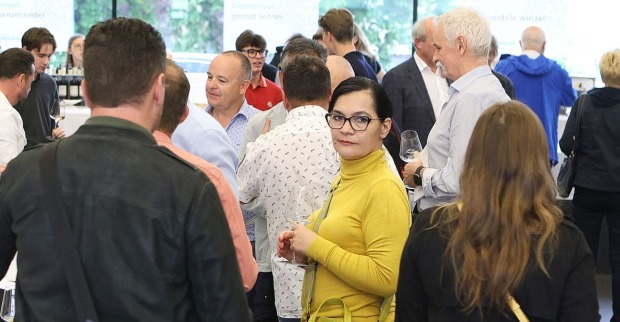 Eine Gruppe von Menschen beim Weinwunder in Linz. In der Bildmitte: Eine Dame mit schwarzen Haaren, gelbem Pullover und Brille hält ein Glas Wein.