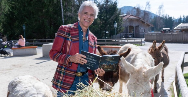 Thomas Brezina mit einem Buch, einem karierten Sakko und umgeben von Eseln.