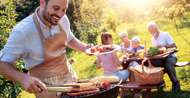 Ein Mann mit schwarzem Vollbart - bekleidet mit einer brauner Schürze grill. In der linken Hand hält er einen Teller mit gegrillten Fleisch und Gemüse, mit der rechten Hand wendet er mithilfe eine Zange einen Maiskolben, der am Grill liegt. Die Sonne scheint, im Hintergrund essen zwei Kinder, eine Frau und ein ältere Herr die gegrillten Speisen.