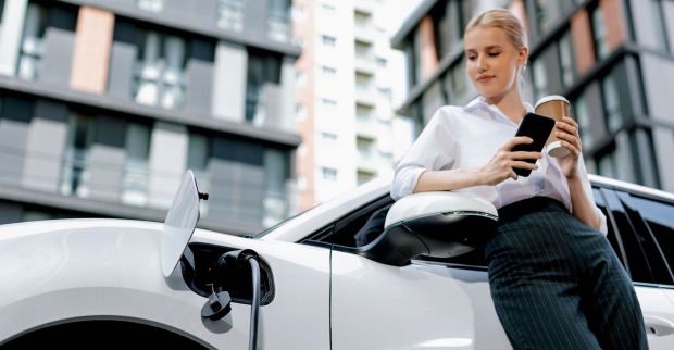Junge Frau mit ihrem Smartphone in der Hand steht gelehnt an ihr Elektroauto | Credit: iStock.com/Ralf Hahn