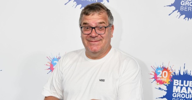 Moderator Elton vor einer Fotowand mit einem weißen T-Shirt.