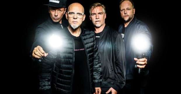 Die Bandmitglieder der "Fantastischen Vier" tragen schwarze Kleidung und blicken ernst in die Kamera. Die zwei Bandmitglieder, die links und rechts stehen, halten auch eine Taschenlampe, die besonders hell leuchtet.