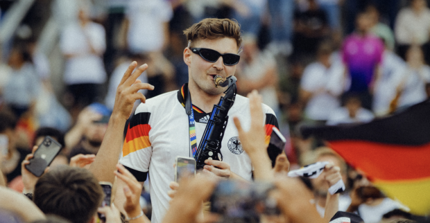 Ein Mann trägt eine Sonnenbrille und spielt ein Saxophon, während ihm alle zujubeln