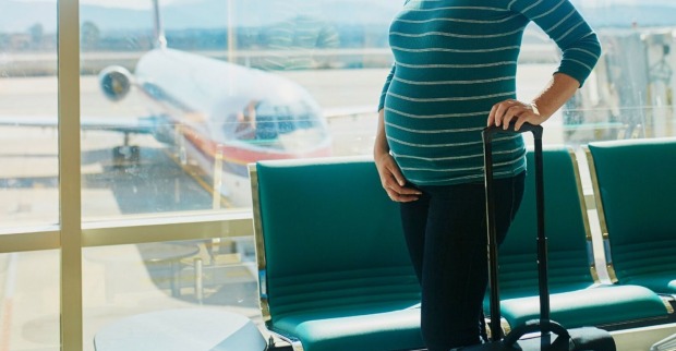 Schwangere Frau am Flughafen die mit einer Hand ihren Bauch hält und mit der anderen Hand einen Koffer schiebt