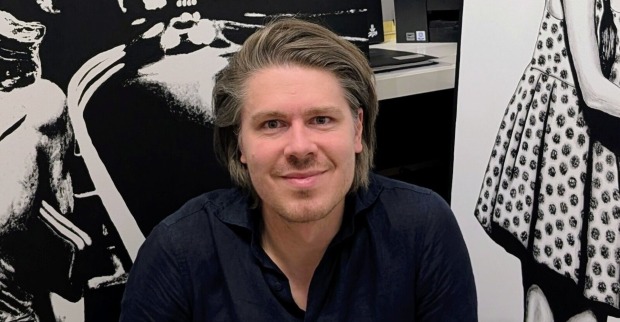 Der Künstler Jürgen Vogel lacht in die Kamera. Er trägt ein blaues Hemd, hat lange braune Haare und einen Drei-Tages-Bart. Im Hintergrund sind Bilder in Schwarz-Weiß zu erkennen.
