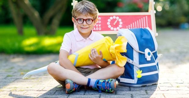 Ein junger Bub - mit kurzer Hose, kurzem Polo und bunten Turnschuhen bekleidet - lacht in die Kamera. Er trägt eine rote Brille und in der Hand hält er eine gelbe Schultüte. Rechts von ihm steht eine blaue Schultasche, hinter ihm eine Kreidetafel auf der Schule zu lesen ist.