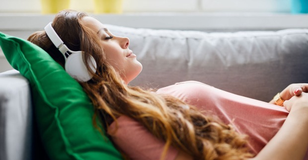 Frau liegt mit Kopfhörern entspannt auf der Couch | Credit: iStock.com/BartekSzewczyk