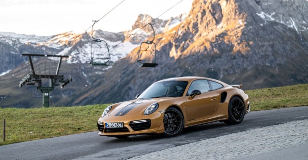 Ein goldener Porsche fährt auf einer schmalen Straße. Im Hintergrund sind schneebedeckte Gipfel und ein Sessellift zu sehen.