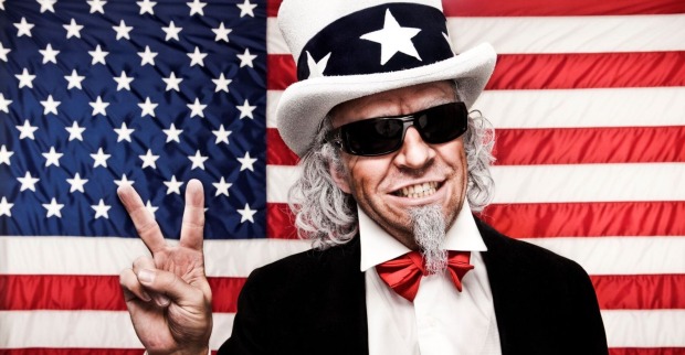 Ein Mann, Symbolfigur Uncle Sam, mit weißem Bart ist zeigt mit seiner rechten Hand das Victory-Zeichen. Er trägt eine schwarze Brille und eine weißen Hut mit schwarzen Band und weißen Sternen. Im Hintergrund ist die Flagge der USA zu sehen.