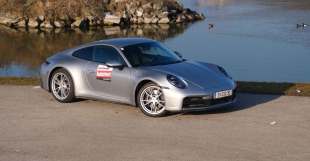 Ein silberner Porsche 911 parkt am Ufer eines Gewässer in dem einige Enten schwimmen. Das Auto leuchtet in der Sonne.