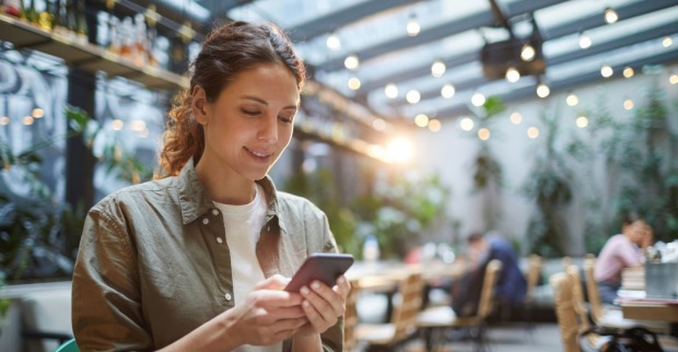 Eine Frau mit braunen Haaren und einem Zopf blickt lächelnd auf ihr Smartphone. Die Frau ist mit einem olivgrünem Hemd und einem weißen T-Shirt bekleidet. Sie sitzt in einem Wintergarten eines Restaurants.