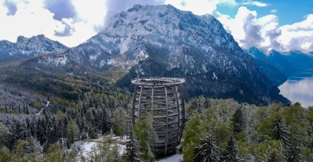 Panoramablick auf den verschneiten Grünberg bei Gmunden mit dem Baumwipfelpfad im Vordergrund und dem Traunstein im Hintergrund. Die umliegenden Wälder sind mit frischem Schnee bedeckt, der Himmel zeigt eine Mischung aus Sonne und dramatischen Wolken.
