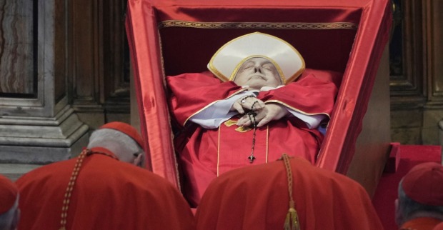Papst Franziskus liegt im roten Gewand mit Mitra und Rosenkranz in einem geöffneten, schräg gestellten Sarg. Vor dem Sarg stehen mehrere Kardinäle in roter Kleidung und verneigen sich.