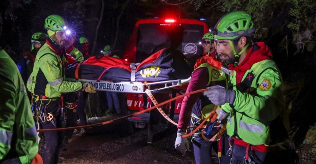 Rettungskräfte in grüner Schutzkleidung und Helmen transportieren bei Nacht eine Person auf einer Trage durch bewaldetes Gelände. Im Hintergrund steht ein Einsatzfahrzeug mit eingeschaltetem Blaulicht.