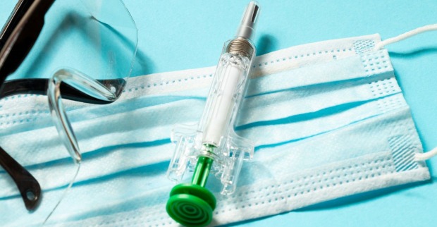 Medizinische Schutzmaske, Schutzbrille und transparente Spritze mit grünem Kolben auf hellblauem Hintergrund – Symbolbild für Pandemie, Impfschutz und Vorsorgemaßnahmen.
