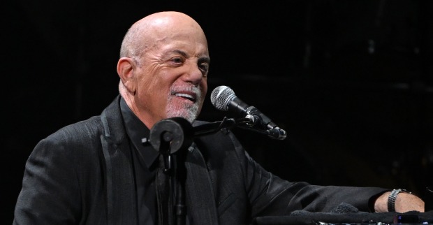 Billy Joel sitzt im dunklen Bühnenlicht an einem Mikrofon und lächelt. Er trägt ein schwarzes Sakko und eine Uhr am Handgelenk.