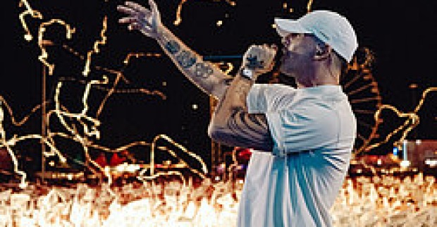 Der Hip-Hop-Musiker RAF Camora performt auf einer Bühne bei Nacht, mit Mikrofon in der Hand und vor einem jubelnden Publikum. Effekte fliegen durch die Luft.