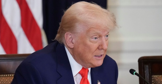 Donald Trump sitzt im Anzug mit roter Krawatte vor einem Mikrofon, im Hintergrund ist eine US-Flagge zu sehen.