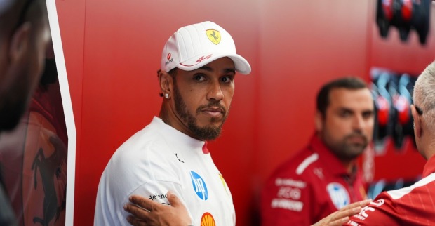 Lewis Hamilton in Ferrari-Teamkleidung steht mit ernster Miene in der Box, umgeben von Teammitgliedern. Deutlich sichtbar: das Ferrari-Logo im Hintergrund.