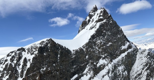Verschneiter, steil aufragender Gipfel des Rimpfischhorns in den Walliser Alpen unter blauem Himmel mit vereinzelten Wolken. Links davon ein schneebedeckter Sattel, rechts steile Felsflanken.