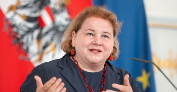 Korinna Schumann bei einer Pressekonferenz, gestikulierend vor den Flaggen Österreichs und der Europäischen Union.