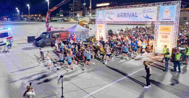 Eine große Gruppe von Läuferinnen und Läufern startet bei einem nächtlichen Lauf-Event auf dem Rollfeld eines Flughafens. Im Vordergrund sind die schnellsten Teilnehmenden bereits losgelaufen. Der Startbereich ist mit einem großen Torbogen mit der Aufschrift „ARRIVAL“ markiert. Rundherum stehen Zuschauende, Ordner in Warnwesten und Fotografen. Links im Bild befinden sich ein Krankenwagen und ein schwarzer Transporter, rechts sind Sponsorenbanner am Starttor zu sehen.