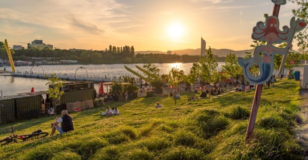 alte Donau im Sommer, Menschen sitzen auf Wiese am Wasser und beobachten den Sonnenuntergang